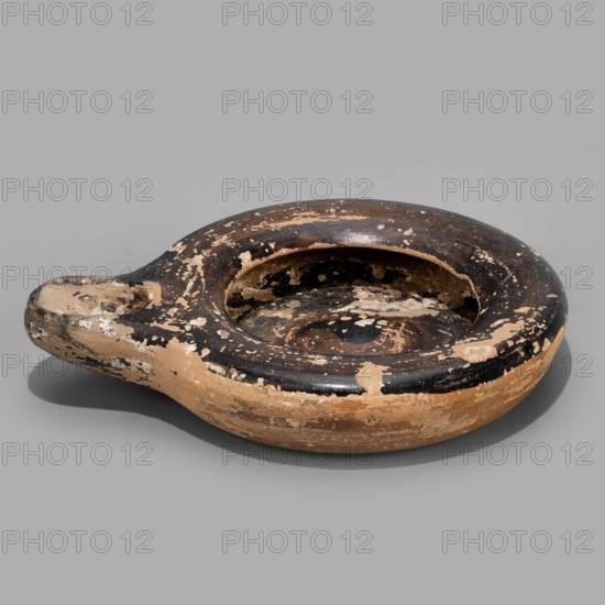 Oil lamp, Reddish, fine-grained clay. Low lamp with broad arms slightly curled inward. Large oil opening, short burner with a large oval wick hole. Slightly concave base. Lustrous fir inside the lamp, on the arms, and on the burner. 5th century BC. Height 1.7 cm; width 9.6 cm., A low, rounded ceramic vessel with a short spout, showing worn and chipped surfaces; the body displays a mix of earthy tan and dark brown to black areas with patches where the outer layer has flaked away, revealing lighter tan beneath; the interior rim and basin show similar wear with mottled dark brown and tan tones; the piece rests on a light gray background and casts a soft shadow.