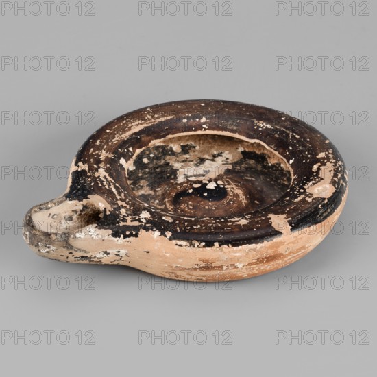 Oil lamp, Tekieli, St. - deposit, Reddish, fine-grained clay. Low lamp with broad arms slightly curled inward. Large oil opening, short burner with a large oval wick hole. Slightly concave base. Lustrous fir inside the lamp, on the arms, and on the burner. 5th century BC. Height 1.7 cm, width 9.6 cm., The image shows a round, shallow ceramic vessel with a short spout-like protrusion, resting on a neutral gray surface. The vessel has a weathered appearance with chipped and flaking areas, revealing a base clay color of warm tan and light beige beneath darker surface layers. The upper rim and interior display a worn dark brown to almost black finish with irregular patches where the darker surface has peeled away, creating mottled contrasts between the dark brown, black, tan, and beige. The vessel's surface texture appears rough in places and smoother in others, and the overall shape is circular with a central depression and an off-center spout.