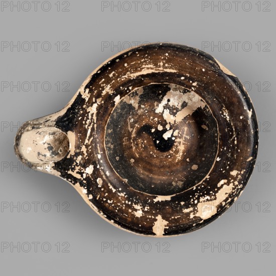 Oil lamp, reddish, fine-grained clay. Low lamp with broad arms slightly curled inward. Large oil opening, short burner with a large oval wick hole. Slightly concave base. Lustrous fir inside the lamp, on the arms, and on the burner. 5th century BC. Height 1.7 cm, width 9.6 cm., A round shallow vessel with a spout-like protrusion, showing worn and chipped surfaces revealing layers of dark brown, black, and beige. The interior has concentric rings of darker brown and black with scattered patches where the top layer has flaked away to show beige underneath. The rim and outer surface display irregular beige spots against a primarily dark brown and black background. The spout area is lighter beige with darker smudges and signs of wear. The background behind the vessel is a uniform gray.
