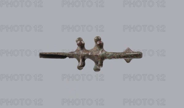Fibula a Drago, One-piece bronze fibula with an arched ring of circular cross-section bearing four pairs of protrusions terminating in spherical bulges. The transition from the ring to the pin is a flat rhombus. Long cylindrical sheath with a slit for the pin. Preservation: ends of the pin and sheath broken off. Date: 950–750 BC 4th quarter 8th–7th century BC. Finding place: Cumae . Type: fibula. Height 3.2 cm; width 6.5 cm., A small metallic object with a rough, irregular surface suspended against a plain light gray background; the object has a central horizontal bar with a few rounded protrusions and bulbous knobs along its top and bottom, and a thin flat extension on one side with a slightly flared end; visible colors include shades of brown, green, and dark gray with areas of darker patina.