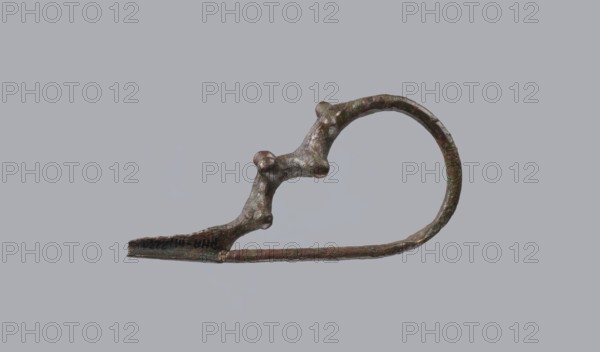 Fibula a Drago, One-piece bronze fibula with an arched ring of circular cross-section bearing four pairs of protrusions terminating in spherical bulges. The transition from the ring to the pin is a flat rhombus. Long cylindrical sheath with a slit for the pin. Preservation: ends of the pin and the sheath are broken off. Dated 950–750 BC 4th quarter 8th–7th century BC. Probably from Cumae. Fibula; height 3.2 cm, width 6.5 cm., A metal object with a curved loop and an elongated straight section, showing an irregular, bumpy surface with rounded protrusions where the curved and straight parts meet; the object appears mottled with greenish and brownish tones and areas of darker brown and black, set against a uniform light gray background.