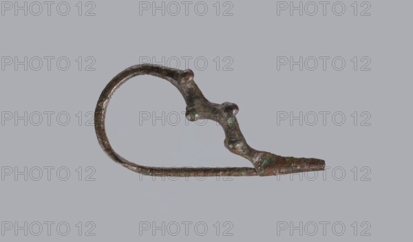 Fibula a Drago, One-piece bronze fibula, with an arched ring of circular cross-section bearing 4 pairs of protrusions terminating in spherical bulges. The transition from the ring to the pin is a flat rhombus. Long cylindrical sheath with a slit for the pin. Preservation: ends of the pin and sheath are broken off. Date: 950–750 BC / 4th quarter 8th–7th century BC. Probable findspot: Cumae. Type: fibula. Dimensions: height 3.2 cm, width 6.5 cm., A single irregular metal object with a looped shape and a protruding tapered end, showing a rough, pitted surface and several rounded bumps along one side; the object displays a mix of brown, green, and dark bronze tones against a plain light gray background.