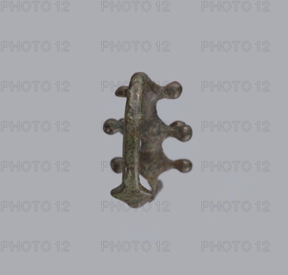 Fibula a Drago, One-piece bronze fibula with an arched ring of circular cross-section bearing four pairs of protrusions terminating in spherical bulges. The transition from the ring to the pin is a flat rhombus. Long cylindrical sheath with a slit for the pin. Preservation: ends of the pin and sheath are broken off. Dated 950–750 BC 4th quarter 8th–7th century BC. Possibly from Cumae. Fibula; height 3.2 cm, width 6.5 cm., A single metallic object with a rough, corroded surface sits against a pale gray background; the object shows a curved central band with several rounded protrusions along one side and a flattened arrowhead-like end, with colors including green, brown, and hints of dark gray.