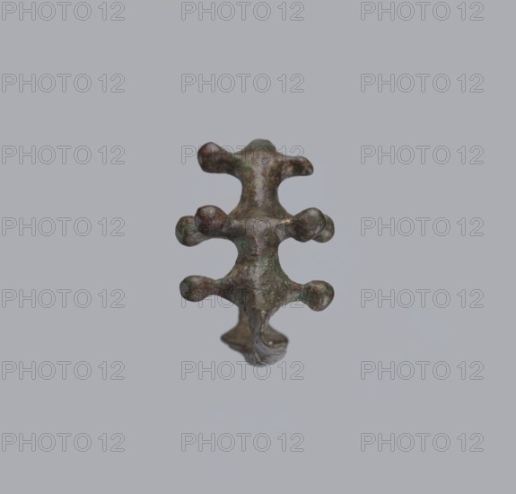 Fibula a Drago, One-piece bronze fibula with an arched ring of circular cross-section bearing four pairs of protrusions terminating in spherical bulges. The transition from the ring to the pin is a flat rhombus. Long cylindrical sheath with a slit for the pin. Ends of the pin and sheath are broken off. Dated 950–750 BC 4th quarter 8th–7th century BC. Findspot: Cumae . Fibula; height 3.2 cm, width 6.5 cm., A small sculptural object with an irregular, organic form floating against a pale gray background; the object has a vertical symmetry with a central spine and several rounded protrusions on either side, and its surface shows a mix of greenish patina, brown, and darker bronze tones with textured, slightly rough areas and subtle highlights.