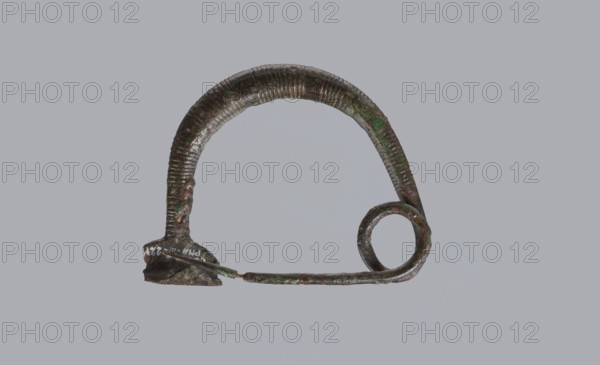 Arched fibula, One-piece fibula with an arched bow made of a round bar, decorated with groups of circumferential notches. A 1.5-turn loop at the transition from the bow to the pin. Condition: sheath damaged. 9th–8th century BC. Southern Italy, possibly Cumae. Bronze. Height 3.2 cm, width 4.2 cm. Fibula ad arco semplice., An aged metal object with a curved arch shape and a spiral loop at one end, showing a textured ribbed surface and irregular corrosion; the metal displays greenish patina and brownish tones against a plain light gray background, with a thin straight rod extending from the base toward the spiral and a small thicker base piece at the opposite end.
