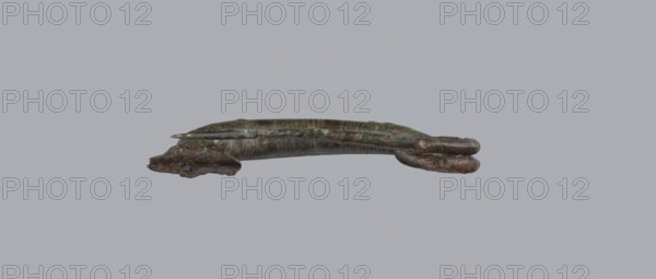 Arched fibula. One-piece fibula with an arched bow made of a round bar, decorated with groups of circumferential notches. A 1.5-turn loop at the transition from the bow to the pin. Condition: sheath damaged. Bronze. 9th–8th century BC. Southern Italy, possibly Cumae. Height 3.2 cm, width 4.2 cm., A small curved metallic object with a weathered, patinated surface in shades of green, brown, and dark gray, appearing slightly corroded and rough in texture; the object has a thin rod or pin resting along its length and rounded, irregular ends with layered or folded metal features, all shown against a uniform light gray background.