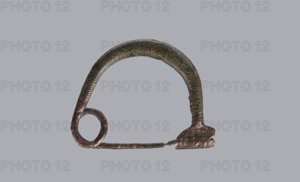 Arched fibula, one-piece ad arco semplice fibula with an arched bow made of a round bronze bar, decorated with groups of circumferential notches. A 1.5-turn loop at the transition from the bow to the pin. Sheath damaged. 9th–8th century BC. Southern Italy, possibly Cumae. Height 3.2 cm, width 4.2 cm., A roughly semicircular metallic object with a textured, ribbed surface and a patina showing green and brown tones, attached on one end to a rounded loop and on the other to an irregular, slightly flattened mount; a thin, straight rod extends between the loop and the mount. The background is a flat, uniform gray.