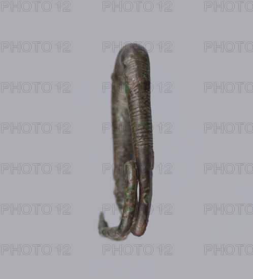 Arched fibula. One-piece fibula with an arched bow made of a round bar, decorated with groups of circumferential notches. A 1.5-turn loop at the transition from the bow to the pin. Condition: sheath damaged. Bronze. 9th–8th century BC. Southern Italy, possibly Cumae. Height 3.2 cm, width 4.2 cm., A single curved metal object suspended against a plain light gray background, showing a textured surface with ridges and a patina of green and brown tones, with a few darker and lighter areas and a slight metallic sheen.