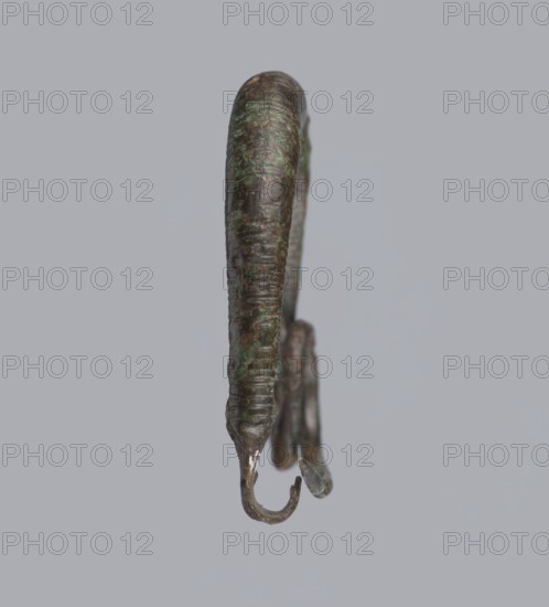 Arched fibula, one-piece fibula with an arched bow made of a round bar, decorated with groups of circumferential notches. A 1.5-turn loop at the transition from the bow to the pin. Condition: sheath damaged. 9th–8th century BC. Southern Italy, Cumae . Fibula, height 3.2 cm, width 4.2 cm., A close-up view of a curved metal object suspended against a plain light gray background, showing a weathered surface with dark brown and greenish patina; the object tapers and has a rounded upper section and a hooked lower end with a smaller hooked fragment beside it, both showing rough texture and mottled areas of green and darker brown.