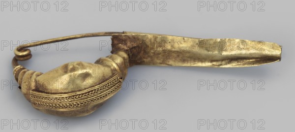 Leech fibula with an elongated stem. The swollen, profiled ring at the ends is decorated with a long strip of filigree in the form of figure eights. The spring has 1 1/2 coils. 2nd half of the 6th century BC, Etruria. Jewelry fibula; height 1.2 cm, width 3 cm, depth 0.6 cm, weight 1 g., A close-up of a metallic object with a curved form, showing a rounded bulbous end attached to a flattened elongated strip; the bulbous section has decorative braided and looped raised bands and a smooth, somewhat uneven surface, and the strip appears crumpled and folded. The object rests on a smooth, neutral background. Visible colors include gold tones on the metal and pale gray in the background.