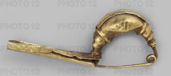 Leech fibula with an elongated stem. Swollen, profiled ring at the ends decorated with a long strip of filigree in the form of figure eights. Spring with 1½ coils. 2nd half of the 6th century BC, Etruria. Jewelry fibula. Height 1.2 cm, width 3 cm, depth 0.6 cm, weight 1 g., A gold-colored metal object shaped like a curved pin with a curled end and a long flat blade-like section, featuring a rounded, sculpted bulge near the curved end with textured braided detail along its top; the surface shows areas of smooth shine and darker tarnish in gold and brown tones against a pale gray background.