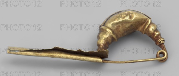 Leech fibula with an elongated stem. The swollen, profiled ring at the ends is decorated with a long strip of filigree in the form of figure eights. The spring has 1 1/2 coils. 2nd half of the 6th century BC, Etruria. Jewelry fibula. Height 1.2 cm; width 3 cm; depth 0.6 cm; weight 1 g., A metallic object photographed on a neutral gray background, showing a curved pin-like form with a looped end and a folded, hollow section; the object has a worn, slightly irregular surface with dents and creases and appears in shades of gold and brass with darker brownish patina in places and small areas of muted gray.