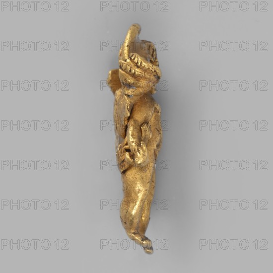 Eros-shaped pendant, Pendant in the shape of a standing Cupid with simplified hands; facial features blurred. Goldsmithery jewelry ornament; necklace pendant. Classified Gallic Scenography; Eros mythology. Black Sea region, 1st half of the 3rd century BC. Height 2 cm, width 1.1 cm, depth 0.4 cm, weight 4 g., A small sculpted figure shown in profile against a pale gray background, rendered in a metallic gold color with areas of darker patina; the figure appears to have a rounded head with textured hair or a headdress, a slightly bent arm with a hand positioned near the torso, and a slender elongated body with a bent knee and a curled foot; the surface shows uneven texture and small marks suggesting wear.