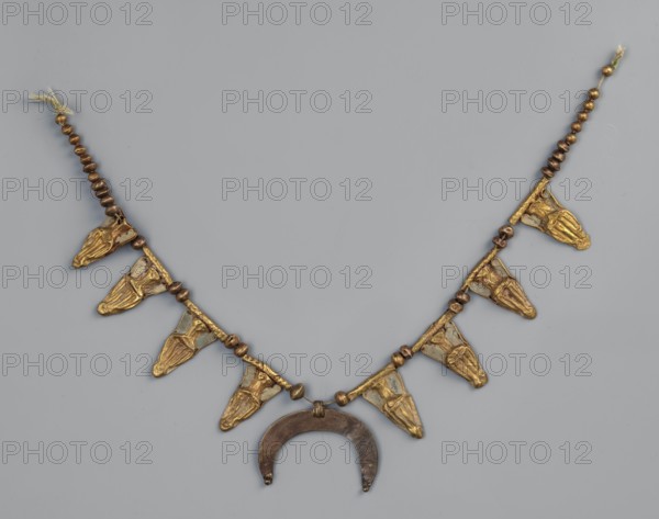Necklace with eight amphorae and a crescent, The necklace consists of eight gold amphorae; the surface between the bodies and the necks of the amphorae is filled with white enamel. Each amphora is attached to a fluted tube. Two gold beads are placed between the amphorae. At the ends of the necklace are 10 beads on one side and 11 beads on the other. In the center hangs a flat downward-facing crescent moon, made of a different, more reddish gold. 3rd century BC possibly late 4th–3rd century BC, Greece, jewelry, weight 12 g., A beaded necklace displayed against a light gray background with a central dark crescent-shaped pendant and a string of small round beads in brown and gold tones leading to a series of triangular gold-colored metal pendants with raised textured details evenly spaced along the lower arc; the triangular pendants have small areas that appear slightly darker or greenish, and the beads vary in metallic brown and muted gold hues, with short tasseled fiber ends at the ends...