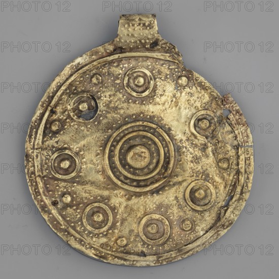 Bulla – medallion, round bulla decorated with concentrically arranged repoussé points; gold, weight 8 g; 3rd quarter of the 8th century BC; Etruria; jewelry, medallion/pendant; goldsmithing., A round metal disc with a tab at the top rests against a flat background; the disc shows a textured, uneven surface with concentric raised rings at the center and several raised circular bosses arranged around it, some of which appear dented or worn. The metal displays a mix of gold and brown tones with darker brown spots and patches of wear, and there are areas where the edge is bent or slightly broken. The background is a solid gray color.