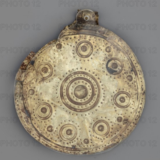 Bulla – medallion, Round bulla decorated with concentrically arranged repoussé points. Gold, pendant/jewelry, weight 8 g. 3rd quarter of the 8th century BC, Etruria., A circular metal object with a worn, mottled surface in shades of gold, brass, brown, and pale green, featuring concentric raised rings and a central rounded boss, surrounded by several smaller raised circular motifs and patterns of small punched dots; the rim is slightly irregular and shows darker brown corrosion and darker spots, and a small tab-like extension projects from the top with varied brownish discoloration; the background is a flat gray.