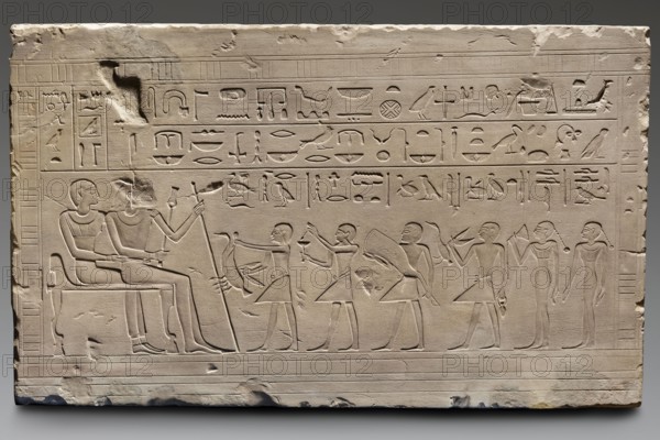 Copy of the Sabni Stele, creator unknown, Sabni ca. 2278–2184 BCE, reliefs; offering iconography; offerings iconography; sepulchral funerary architecture; see original; 20th century; Warsaw; copy; height 53 cm; width 88 cm; depth 10 cm; weight 95 kg, A carved stone relief panel in pale beige with shallow incised figures and symbols covering most of its surface, showing a seated pair on the left with one figure holding a long staff and the other touching the first, several standing figures to the right offering objects and holding items such as vessels and a curved piece, and a group of standing figures at the far right, all rendered in outline with smooth surfaces and slight wear. The upper portion contains rows of repeated carved pictorial signs carved in horizontal bands. The lower edge shows parallel incised lines. The panel has chips, abrasions, and rough edges with darker areas where material is missing, and subtle shadowing emphasizes the carved lines. Colors visible include pale...