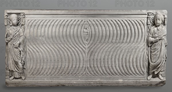 Sarcophagus with a depiction of a married couple and a shepherd, unknown Roman workshop. A large Roman sarcophagus with strigilla S-shaped grooves forming a central medallion, decorated with a standing, thoughtful shepherd leaning on a shepherd's staff pedum. On the front sides are figures presumed to be the deceased, standing against draped fabrics parapetasmata: on the left, a woman in a tunic and long cloak palla holding a scroll volumen in her left hand; on the right, a man in tunic and toga with a contabulatio specially wrapped hem shaped like a belt, holding a scroll in his left hand and wearing mullei leather military boots. Both frontal figures have incompletely finished heads. The side walls feature reliefs of a seated griffin with a lion's head. The left wall has an opening to the interior near its lower left corner; the lower left corner of the right wall is missing. The rear wall is rather carelessly worked. Quite a few abrasions and damage, particularly to the corners and ...