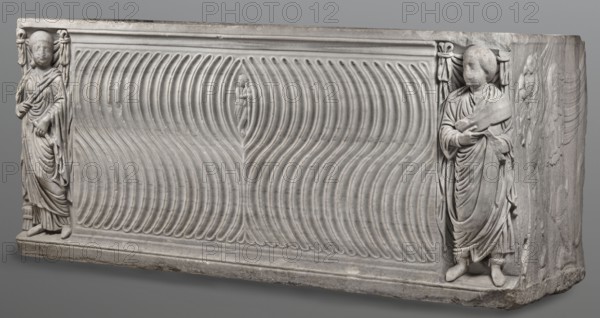 Sarcophagus with a depiction of a married couple and a shepherd, unknown Roman workshop. A large Roman sarcophagus with strigilla S-shaped grooves converging toward the center forming a central medallion, decorated with a standing, thoughtful shepherd leaning casually on a shepherd's staff pedum. On the front sides are figures likely representing the deceased, standing against draped fabrics parapetasmata. Left: a woman in a tunic and long cloak palla, holding a scroll volumen in her left hand. Right: a man in a tunic and toga with a contabulatio specially wrapped hem shaped like a belt, holding a scroll in his left hand and wearing mullei leather military boots. Both frontal figures have incompletely finished heads. The side walls are decorated with reliefs of a seated griffin with a lion's head; the left wall near its lower left corner has an opening into the interior; the lower left corner of the right wall is missing. The rear wall is rather carelessly worked. Quite a few abrasions...