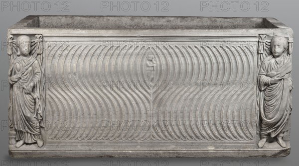 Sarcophagus with a depiction of a married couple and a shepherd, unknown Roman workshop. A large Roman sarcophagus with strigilla S-shaped grooves forming a central medallion, decorated with a standing, thoughtful shepherd leaning on a shepherd's staff pedum. On the sides of the front are figures presumed to be the deceased, standing against draped fabrics parapetasmata: on the left, a woman in a tunic and long cloak palla holding a scroll volumen in her left hand; on the right, a man in a tunic and toga with contabulatio a specially wrapped hem, holding a scroll in his left hand and wearing calcei or military-style leather boots. Both frontal figures have incompletely finished heads. The side walls show reliefs of a seated griffin with a lion's head; the left wall near its lower left corner has an opening to the interior. The lower left corner of the right wall is missing and the rear wall is rather carelessly worked. Considerable abrasions and damage, particularly to the corners and ...