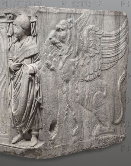 Sarcophagus with a depiction of a married couple and a shepherd, unknown Roman workshop. A large Roman sarcophagus with strigilla S-shaped grooves forming a central medallion, decorated with a standing, thoughtful shepherd leaning on a shepherd's staff pedum. On the front sides are figures presumed to be the deceased, standing against draped fabrics parapetasmata: on the left, a woman in a tunic and long cloak palla holding a scroll volumen in her left hand; on the right, a man in a tunic and toga with a contabulatio specially wrapped hem shaped like a belt, holding a scroll in his left hand and wearing calcei or military-style leather boots. Both frontal figures have incompletely finished heads. The side walls show reliefs of a seated griffin with a lion's head; the left wall near its lower left corner has an opening into the interior, and the lower left corner of the right wall is missing. The rear wall is rather carelessly worked. Noticeable abrasions and damage, particularly to cor...