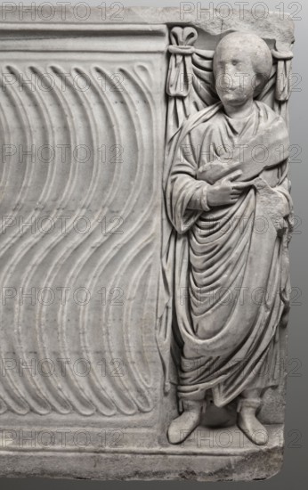 Sarcophagus with a depiction of a married couple and a shepherd, unknown Roman workshop. A large Roman sarcophagus with strigilla S-shaped grooves converging toward the center forming a central medallion, decorated with a standing, thoughtful shepherd leaning on a shepherd's staff pedum. On the front sides are figures presumed to be the deceased, standing before draped fabrics parapetasmata: on the left, a woman in a tunic and long cloak palla holding a scroll volumen in her left hand; on the right, a man in a tunic and toga with contabulatio specially wrapped hem, holding a scroll in his left hand and wearing mullei leather military boots. Both frontal figures have incompletely finished heads. The side walls show reliefs of a seated griffin with a lion's head; the left wall near its lower left corner has an opening into the interior, and the lower left corner of the right wall is missing. The rear wall is rather carelessly worked. Considerable abrasions and damage, particularly at the...