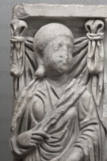 Sarcophagus with a depiction of a married couple and a shepherd, unknown Roman workshop. A large Roman sarcophagus with strigilla S-shaped grooves converging toward a central medallion, decorated with a standing, thoughtful shepherd leaning on a pedum shepherd's staff. The front sides show presumed depictions of the deceased against draped fabrics parapetasmata: on the left, a woman in a tunic and palla holding a scroll volumen in her left hand; on the right, a man in tunic and toga with a contabulatio specially wrapped hem holding a scroll in his left hand and wearing calcei leather military boots. Both frontal figures have incompletely finished heads. The side walls bear reliefs of a seated griffin with a lion's head; the left wall near its lower left corner has an opening to the interior; the lower left corner of the right wall is missing. The rear wall is rather carelessly worked. Considerable abrasions and damage, particularly to the corners and figures on both sides of the front ...