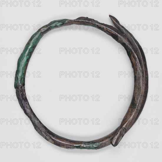 Bracelet, unknown northern Mesopotamian workshop. Small round bracelet made of a single coil of rod, rounded on the outside and conical on the inside, the ends overlap by one-third of the bracelet's circumference. 1st half of the 3rd millennium BC; Early Jazira III period, Northern Mesopotamia; found at Tell Rad Shakra Northeastern Syria. Height 5 cm., A roughly circular metal ring with an irregular, twisted shape, showing a patina of green and areas of dark brown and black; the surface is textured and uneven with a few rough, corroded patches and a slightly pointed overlapping end where the metal layers meet, set against a plain light gray background.