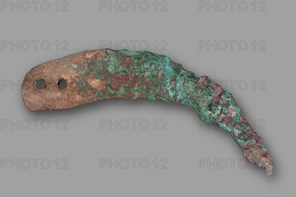 Sickle blade, unknown Northern Mesopotamian workshop. The sickle blade is arched, tipped with a pointed tip; the other end is wide, rounded, with two round holes cut for rivets to attach a handle. On the inside there is a bevel on the cutting edge. 2nd half of the 3rd millennium BC; Early Jazira IV period Akkadian period, ca. 2350–ca. 2200 BC. From Tell Rad Shakra North-Eastern Syria, Northern Mesopotamia Khabur basin. Tool. Weight: 44 g., An irregular, curved metallic object with a rough, corroded surface lying against a plain gray background, showing a mix of brown, green, teal, and reddish areas; the object has a thicker flat end with two circular holes and a thinner, jagged tapering end with uneven textured protrusions, and the surface displays mottled patches and rough pitting across its length.