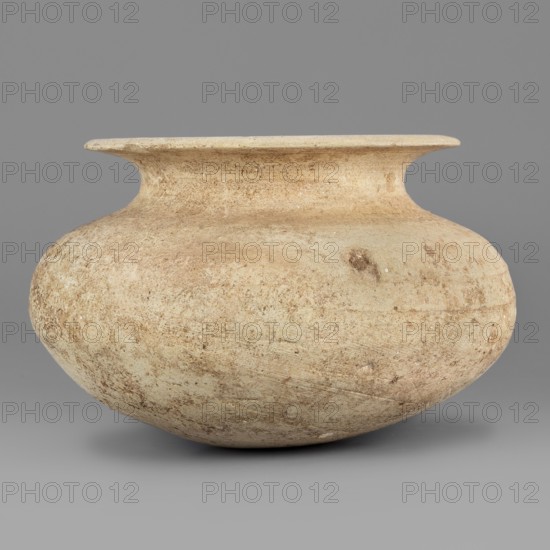 Vessel, unknown northern Mesopotamian workshop; viseless vessel for oils and ointments; low vessel with an ellipsoidal belly with a rounded bottom, low neck, and wide upturned rim; 1st half of the 3rd millennium BC, Early Jazira III period, Northern Mesopotamia Khabur Basin, Tell Rad Shakra Northeastern Syria; vessel; height 6.8 cm, width 10.8 cm, weight 200 g., A rounded, wide-mouthed ceramic vessel with a flattened rim and a short neck sits against a plain background; its surface shows a textured, weathered appearance with variations of beige, tan, and light brown, including patches and speckles of darker brown and subtle lighter off-white areas, and a few small surface imperfections.