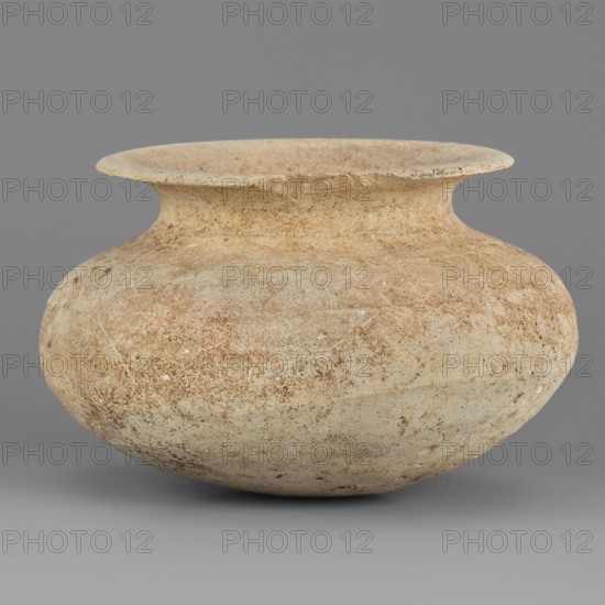 Vessel, unknown northern Mesopotamian workshop; viseless vessel for oils and ointments; low vessel with an ellipsoidal belly with a rounded bottom, low neck, and wide, upturned rim. 1st half of the 3rd millennium BC; Early Jazira III period, Northern Mesopotamia Khabur Basin, Tell Rad Shakra Northeastern Syria. Height 6.8 cm, width 10.8 cm, weight 200 g., A rounded, wide-mouthed ceramic vessel sits against a plain gray background; its surface shows a weathered, textured appearance with mottled tones of beige, tan, light brown, and hints of pale reddish and off-white, and the rim flares outward with a slightly uneven edge while the body bulges into a bulbous shape and narrows slightly toward the neck.