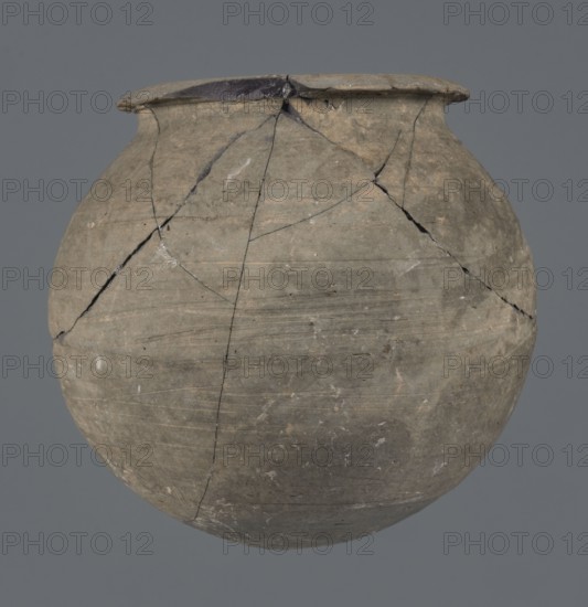 Vessel, unknown Northern Mesopotamian workshop, Early Jazira III period ca. 2600–ca. 2350 BC, metallic ware; spherical, vice-free vessel for oils and ointments. Low vessel with a spherical belly, rounded bottom and low, wide neck with a narrow, upturned spout. The vessel was carefully smoothed and fired in a reduction atmosphere. Syria, Northern Mesopotamia Khabur basin, Tell Rad Shakra. Height 9.4 cm; width 9.7 cm., A rounded clay vessel with a wide rim sits against a gray background, showing a weathered surface in shades of gray, brown, and muted tan with visible fine horizontal striations and areas of lighter and darker discoloration; the vessel has several hairline cracks and repaired joins radiating from the upper portion and rim, with small chips and rough edges around the lip, and a few pale flecks or spots scattered across its body.