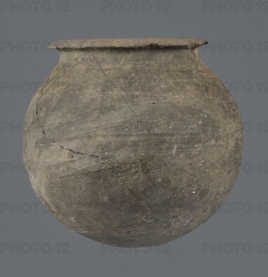 Vessel, unknown Northern Mesopotamian workshop, Early Jazira III period ca. 2600–ca. 2350 BC, metallic ware, spherical vessel for oils and ointments, low vessel with a spherical belly, rounded bottom and low, wide neck with a narrow, upturned spout. The vessel was carefully smoothed and fired in a reduction atmosphere. Syria Tell Rad Shakra, Khabur basin, Northeastern Syria, Northern Mesopotamia place of production, height 9.4 cm, width 9.7 cm., A rounded, weathered clay vessel with a wide rim and bulbous body sits against a plain gray background; the vessel shows a rough surface texture with visible cracks and hairline fractures, areas of uneven wear, and small pits and flecks; the colors visible include muted gray, brown, beige, and hints of darker gray tones across the pot.