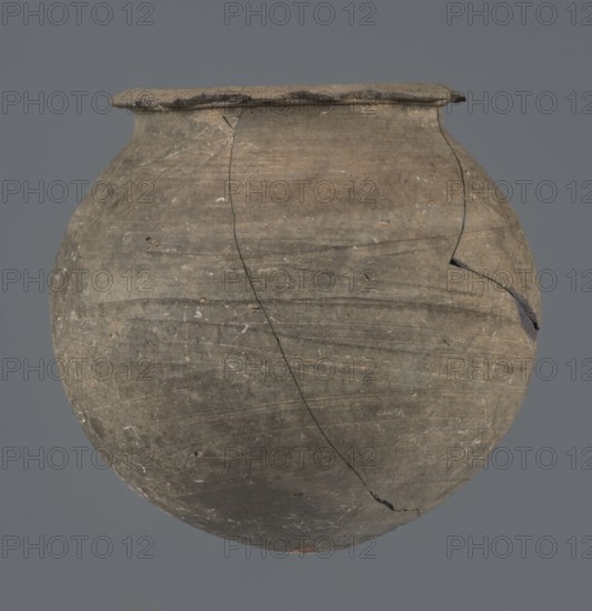 Vessel, unknown Northern Mesopotamian workshop, Early Jazira III period ca. 2600–ca. 2350 BC, metallic ware, spherical vessel for oils and ointments, low vessel with a spherical belly, rounded bottom and low, wide neck with a narrow, upturned spout. The vessel was carefully smoothed and fired in a reduction atmosphere. Syria Tell Rad Shakra, Khabur basin, Northern Mesopotamia place of production, height 9.4 cm, width 9.7 cm., The image shows a round, weathered clay pot with a wide rim, displayed against a plain gray background; the pot's surface is mostly muted brown and gray with darker gray and slightly greenish tints, visible hairline cracks and chips, areas of lighter beige and off-white speckling, and a few darker fractured edges.
