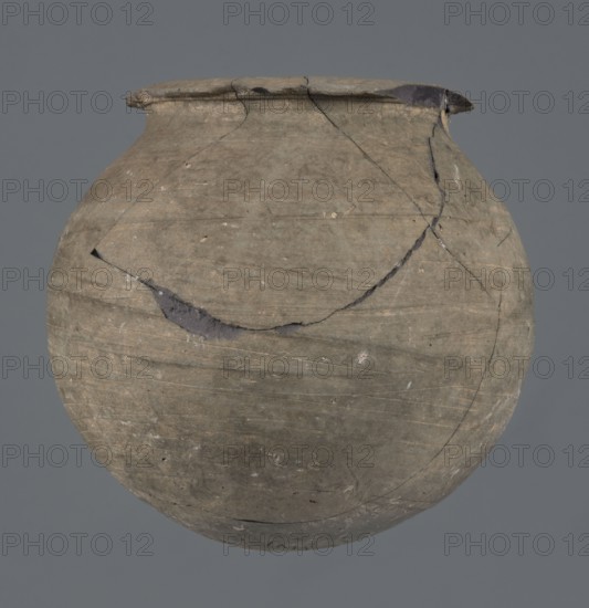 Vessel, unknown Northern Mesopotamian workshop, Early Jazira III period ca. 2600–ca. 2350 BC, metallic ware; spherical, vice-free vessel for oils and ointments. Low vessel with a spherical belly, rounded bottom and low, wide neck with a narrow, upturned spout. The vessel was carefully smoothed and fired in a reduction atmosphere. Ca. 2600–ca. 2350 BC; Northern Mesopotamia Khabur basin, Tell Rad Shakra Northeastern Syria. Height 9.4 cm, width 9.7 cm., A round, worn clay vessel with a narrow rim and a few visible cracks and chips, showing irregular repair lines and rough surface texture; the colors visible are gray, brown, and dark gray, set against a solid muted gray background.
