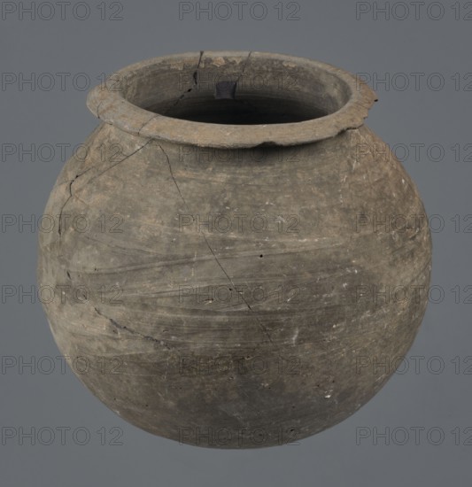 Vessel, unknown Northern Mesopotamian workshop, Early Jazira III period ca. 2600–ca. 2350 BC, metallic ware; spherical, vice-free vessel for oils and ointments. Low vessel with a spherical belly, rounded bottom and low, wide neck with a narrow, upturned spout. The vessel was carefully smoothed and fired in a reduction atmosphere. ca. 2600–ca. 2350 BC; Syria, Northern Mesopotamia Khabur basin, Tell Rad Shakra. Vessel, height 9.4 cm, width 9.7 cm., A round clay vessel with a wide rim and an open top sits against a plain gray background, showing a weathered surface with visible cracks and repaired seams, textured horizontal striations, and small chips and pitted spots; the vessel displays shades of brown, gray, taupe, and darker earthy tones with lighter flecks and a darker interior.