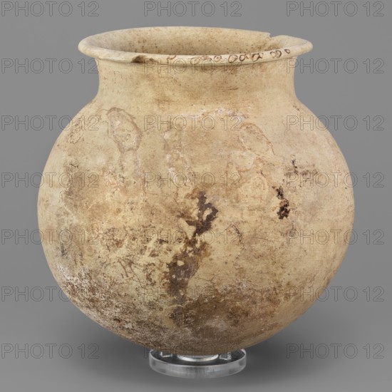 Vessel with a spherical belly, unknown northern Mesopotamian workshop; clay, spherical vessel with a low cylindrical neck terminating in a thickened outwardly curled rim, rounded bottom; 1st half of 3000 BC; Early Jazira III period Early Dynastic III; ca. 2600–ca. 2350 BC; Northern Mesopotamia Khabur basin, Tell Rad Shakra North-Eastern Syria; vessel; height 15.6 cm, width 15.8 cm, weight 600 g., A rounded earthenware vessel with a wide flared rim sits against a gray background, showing a surface of various beige and tan tones with areas of darker brown discoloration and lighter cream patches; the pot has a weathered, slightly rough texture with irregular marks and stains, and rests on a small clear circular stand that appears translucent.