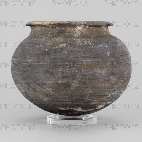Vessel, unknown northern Mesopotamian workshop; viceless vessels; metallic ware; spherical vessel; vessel for oils and ointments; Low vessel with a spherical belly, rounded bottom, low wide neck with a narrow, upturned spout. 1st half of the 3rd millennium BC; Early Jazira III period, Northern Mesopotamia Khabur Basin, Tell Rad Shakra Northeastern Syria. Vessel; height 8.3 cm, width 10.6 cm, weight 220 g., A rounded ceramic vessel with a wide neck and flared rim sits on a clear circular stand; the surface of the vessel shows a weathered, uneven texture with areas of wear and small spots where the finish has flaked, revealing lighter patches; its dominant colors are dark gray and charcoal with streaks and subtle bands of brown, muted tan, and faint whitish marks; the stand is translucent and colorless, and the background is a pale light gray.