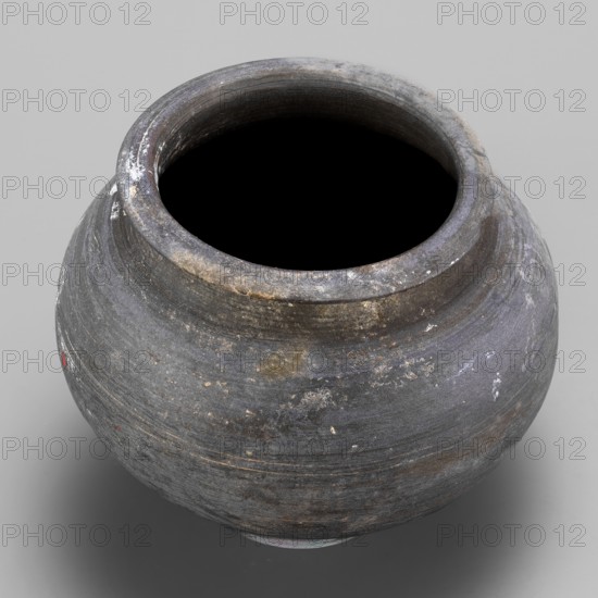 Vessel, unknown northern Mesopotamian workshop; viceless vessel, metallic ware, spherical vessel for oils and ointments; low vessel with a spherical belly, rounded bottom, low wide neck with a narrow, upturned spout. 1st half of the 3rd millennium BC; Early Jazira III period, Northern Mesopotamia Khabur Basin, Tell Rad Shakra Northeastern Syria. Height 8.3 cm, width 10.6 cm, weight 220 g., A rounded ceramic vessel with a wide open mouth sits against a neutral background, showing a weathered surface in shades of gray and charcoal with subtle bluish and brownish tints, areas of lighter whitish residue and small patches of reddish-brown, a few rough spots and fine horizontal striations from its making, and a dark interior visible through the opening.