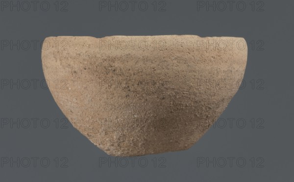 Bowl, unknown Northern Mesopotamian workshop, Early Jazira III period ca. 2600–ca. 2350 BC. Clay vessel tableware. Bowl with a flat, obliquely cut round bottom, gently sloping walls that straighten in the upper part and bend slightly inward, ending in a straight, non-distinctive rim. Before firing, a potter's mark was engraved at the bottom as two intersecting lines of differing lengths: a shorter line parallel to the bottom and a longer line starting at the belly and reaching half the bottom diameter. Origin: Northern Mesopotamia Khabur basin, Tell Rad Shakra, Northeastern Syria., A roughly bowl-shaped object with a coarse, textured surface in shades of tan, beige, and light brown suspended against a flat dark gray background; the rim is uneven and the body shows mottled darker brown speckles and small lighter flecks across its surface.