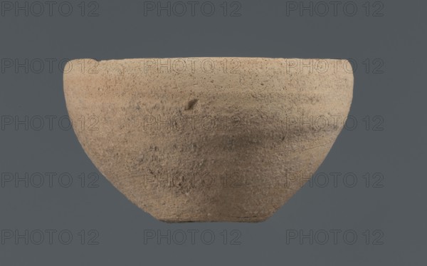 Bowl, unknown Northern Mesopotamian workshop, Early Jazira III period ca. 2600–ca. 2350 BC Early Dynastic III period. Clay vessel: bowl with a flat, obliquely cut, round bottom, gently sloping walls that straighten in the upper part and bend slightly inwards to a straight, non-distinctive rim. Before firing a potter's mark was engraved at the bottom: two intersecting lines of varying lengths — the shorter line runs parallel to the bottom, the longer starts at the belly and reaches half the bottom diameter. Origin: Northern Mesopotamia Khabur basin, Tell Rad Shakra, northeastern Syria., A roughly hemispherical bowl with a coarse, textured surface sits against a solid background; the bowl shows an earthy beige and light brown coloration with subtle darker brown patches and small speckled areas, and the background is a muted grayish blue.