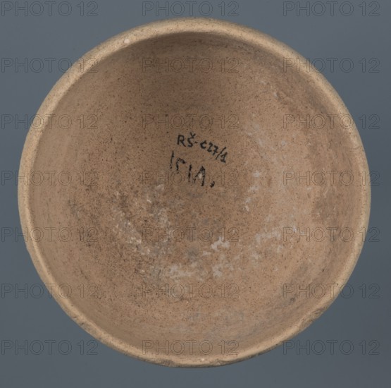 Bowl, unknown Northern Mesopotamian workshop, Early Jazira III period ca. 2600–ca. 2350 BC Early Dynastic III period. Clay vessel: bowl with a flat, obliquely cut round bottom, gently sloping walls that straighten in the upper part and bend slightly inward, ending in a straight, non-distinctive rim. Before firing, a potter's mark was engraved at the bottom as two intersecting lines of varying lengths: a shorter line parallel to the bottom and a longer line starting at the belly and reaching half the bottom diameter. ca. 2600–ca. 2350 BC; northern Mesopotamia Khabur basin, Tell Rad Shakra, northeastern Syria., A round shallow bowl viewed from above with an inner surface showing a rough, sandy texture in shades of light brown and tan with patches of pale whitish residue and darker brown speckling; the rim is slightly thicker and uniform in a beige tone, and the background around the bowl is a muted blue-gray.