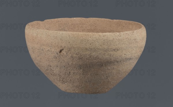 Bowl, unknown Northern Mesopotamian workshop, Early Jazira III period ca. 2600–ca. 2350 BC Early Dynastic III period. Clay vessel: bowl with a flat, obliquely cut round bottom, gently sloping walls that straighten in the upper part and bend slightly inward, ending in a straight, non-distinctive rim. Before firing a potter's mark was engraved at the bottom as two intersecting lines of varying lengths—the shorter running parallel to the bottom, the longer starting at the belly and reaching half the bottom diameter. Origin: Northern Mesopotamia Khabur basin, Tell Rad Shakra, northeastern Syria., A roughly bowl-shaped clay vessel occupies the image, shown against a solid muted dark gray background; the bowl has a coarse, textured surface with visible grain and small pits, and its form is rounded with a slightly uneven rim. The visible colors include light tan, beige, sandy brown, and areas of darker brown and grayish tones, with subtle variations and mottling across the exterior and interi...