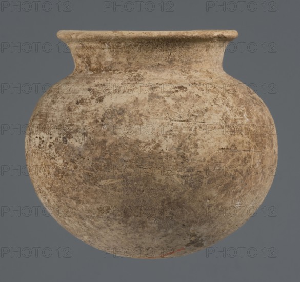 Globular vessel, unknown Northern Mesopotamian workshop; clay; Early Jazira III period ca. 2600–ca. 2350 BC Early Dynastic III; globular vessel with a low, conically flaring neck terminating in a narrow, slightly thickened and everted rim; rounded, slightly flattened bottom; Northern Mesopotamia Khabur basin, Tell Rad Shakra, Syria; height 8.4 cm, width 9.5 cm., A rounded clay vessel with a short, flared neck and a wide lip, shown against a flat gray background; the vessel surface appears textured and weathered with a mix of beige, tan, light brown, and darker brown tones, and a few subtle reddish-orange spots near the lower edge.