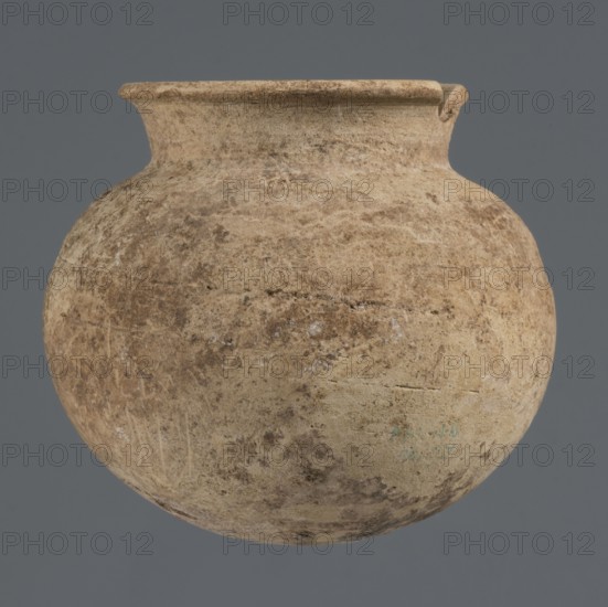Globular vessel, unknown Northern Mesopotamian workshop; clay; Early Jazira III / Early Dynastic III period ca. 2600–ca. 2350 BC. Globular vessel with a low, conically flaring neck terminating in a narrow, slightly thickened and everted rim; rounded, slightly flattened bottom. Origin: Northern Mesopotamia Khabur basin, Tell Rad Shakra, Al-Jazira Syria/Iraq. Vessel; height 8.4 cm, width 9.5 cm., A rounded, bulbous vessel with a short, flaring neck and a slightly thickened rim sits against a solid background; the surface of the vessel shows a weathered, uneven texture with mottled patches and speckling in shades of beige, tan, brown, and hints of cream, and there are areas of lighter and darker discoloration and subtle surface abrasions.