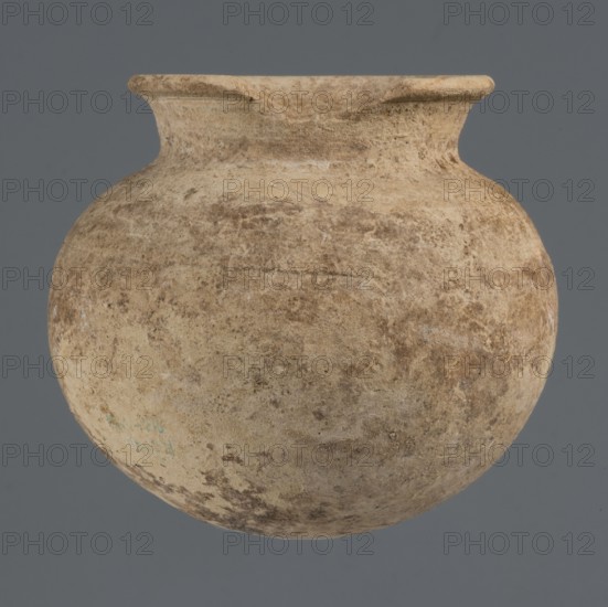 Globular vessel, unknown Northern Mesopotamian workshop; clay; Early Jazira III period ca. 2600–ca. 2350 BC; Early Dynastic III period. Globular vessel with a low, conically flaring neck terminating in a narrow, slightly thickened and everted rim. Rounded, slightly flattened bottom. Origin: Northern Mesopotamia Khabur basin, Tell Rad Shakra, Syria. Height 8.4 cm; width 9.5 cm., A rounded clay vessel with a wide bulbous body and a short neck topped by a flared rim fills the frame, its surface showing a weathered, slightly rough texture with irregular patches and mottling. The vessel displays a palette of beige, tan, and brown tones with areas of darker browning and subtle lighter cream highlights, and there are hints of muted greenish spots toward one side. The background behind the vessel is a flat, even gray.
