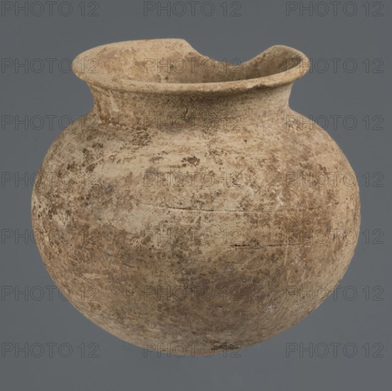 Globular vessel, unknown Northern Mesopotamian workshop, clay, Early Jazira III period ca. 2600–ca. 2350 BC; Early Dynastic III period. Globular vessel with a low, conically flaring neck terminating in a narrow, slightly thickened and everted rim; rounded, slightly flattened bottom. Northern Mesopotamia Khabur basin, Tell Rad Shakra North-eastern Syria, ca. 2600–ca. 2350 BC. Vessel; height 8.4 cm, width 9.5 cm., A round, wide-mouthed clay vessel with a flared rim and an irregular chip on one side, showing a rough, textured surface with mottled tones of beige, tan, brown, and hints of gray, set against a plain muted gray background.