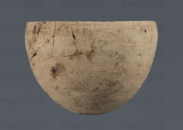 Bowl, unknown northern Mesopotamian workshop, Early Jazira III period ca. 2600–ca. 2350 BC / Early Dynastic III period, clay, bowl with an egg-shaped body, rounded bottom and a straight, non-separated rim slightly bent inwards, ca. 2600–ca. 2350 BC, Northern Mesopotamia Khabur basin, Tell Rad Shakra North-Eastern Syria, Syria; vessel; height 7.8 cm; width 10.7 cm, A light beige rounded clay vessel with a slightly uneven rim and visible surface cracks and repairs sits against a flat muted blue-gray background, showing a textured surface with patches of darker brown discoloration and small pitted marks.