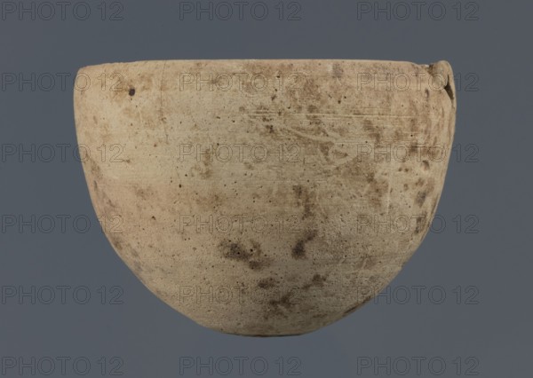 Bowl, unknown northern Mesopotamian workshop, Early Jazira III period ca. 2600–ca. 2350 BC / Early Dynastic III period ca. 2600–ca. 2350 BC. Clay bowl with an egg-shaped body, rounded bottom, and a straight, non-separated rim slightly bent inwards. Northern Mesopotamia Khabur basin; found at Tell Rad Shakra, North-Eastern Syria. Vessel; height 7.8 cm, width 10.7 cm., A rounded, bowl-shaped ceramic object with a rough, weathered surface in beige and off-white tones, showing a mottled pattern of darker brown and tan spots and smudges; the rim is slightly irregular with a small protrusion on one side, and the background is a solid muted gray-blue.