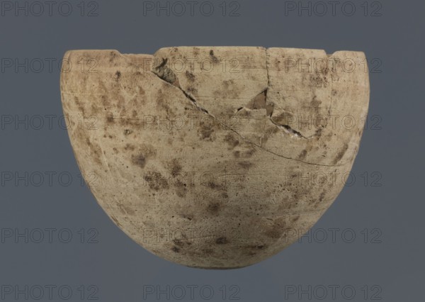 Bowl, unknown northern Mesopotamian workshop, Early Jazira III period ca. 2600–ca. 2350 BC / Early Dynastic III period ca. 2600–ca. 2350 BC. Clay vessel; egg-shaped bowl with a rounded bottom and a straight, non-separated rim slightly bent inward. Northern Mesopotamia Khabur basin; found at Tell Rad Shakra, North-Eastern Syria. Vessel; height 7.8 cm, width 10.7 cm., A rounded ceramic bowl with a chipped rim and visible cracks sits against a solid gray background; the bowl shows a weathered, mottled surface in beige and tan with darker brown spots and smudges, and a few lighter worn areas where the material appears paler.